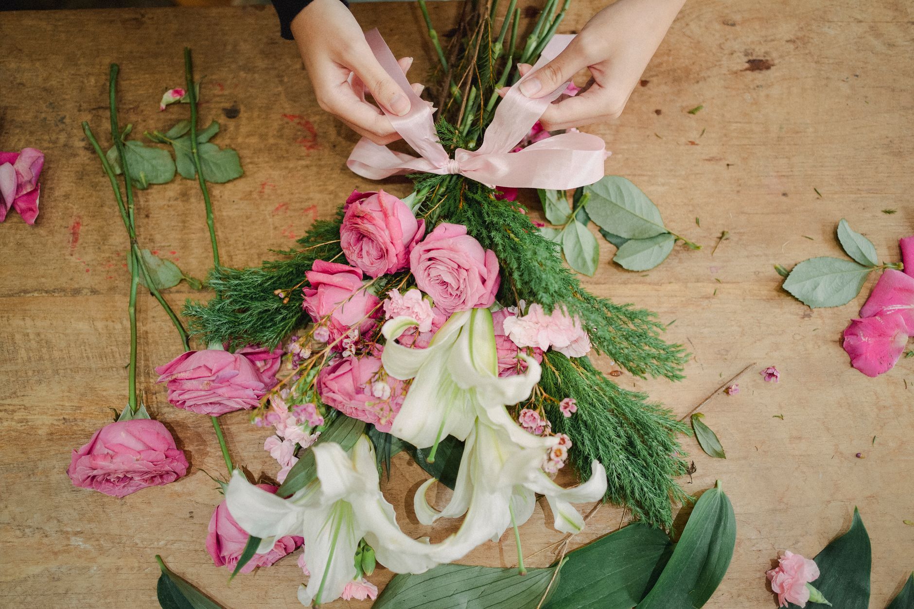 Hands tying a pink ribbon on a bouquet of pink roses, white lilies, and evergreen on a wooden table