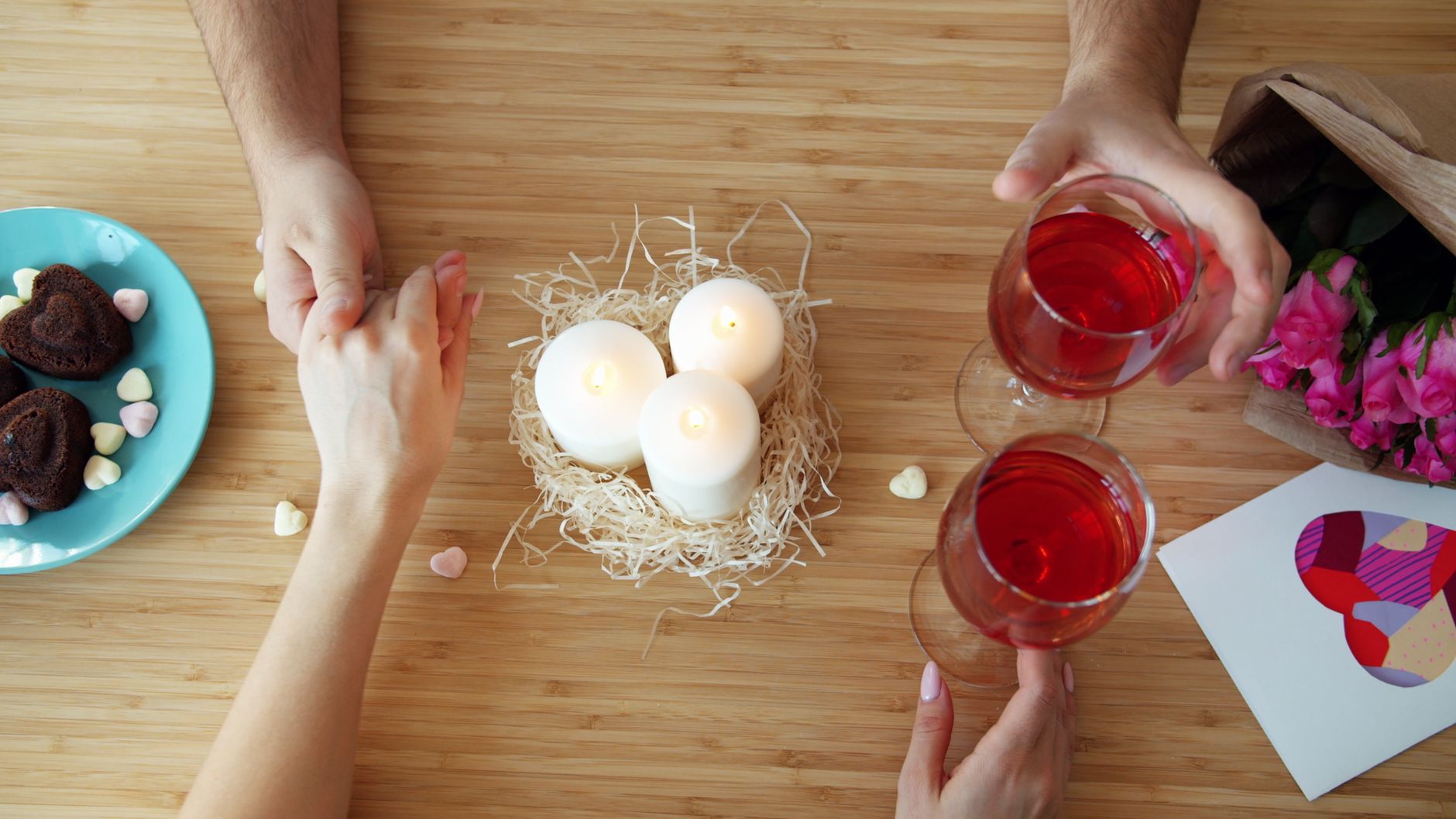 Two people holding hands over a table with candles, red wine, chocolates, and pink flowers