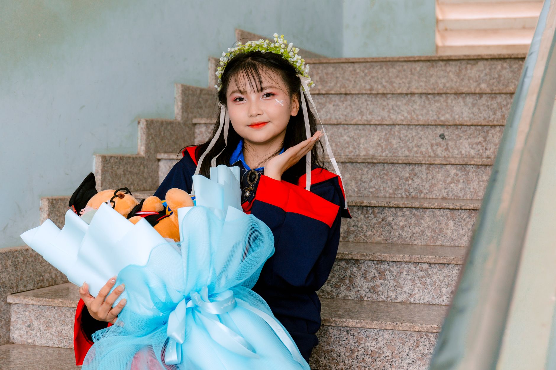 A young graduate in a gown wearing a flower wreath and holding a pale blue bouquet on a staircase