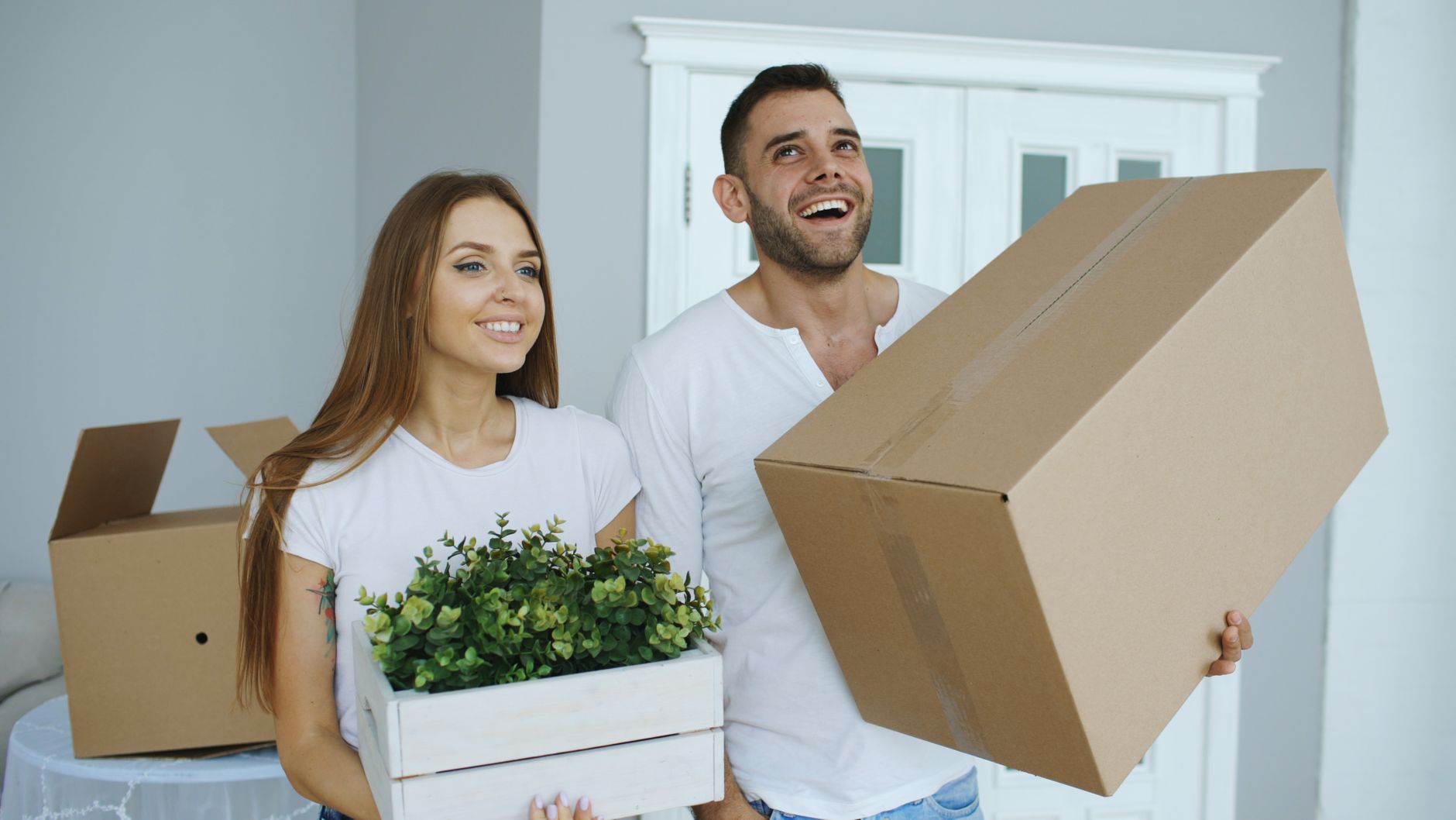 A smiling couple carrying moving boxes and a crate of green plants into a new home