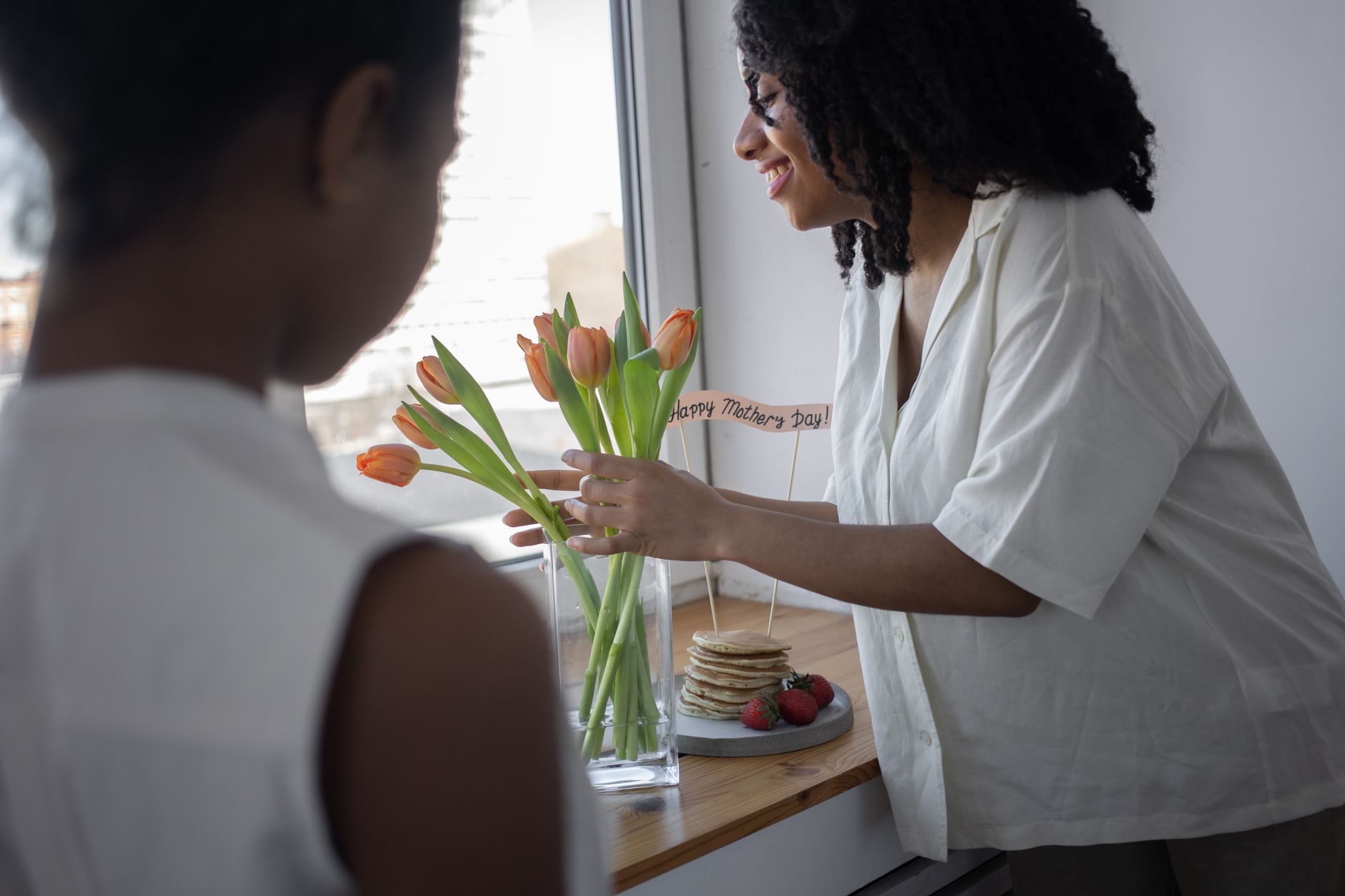 A woman arranging orange tulips in a glass vase beside a stack of pancakes and a Happy Mother&rsquo;s Day sign