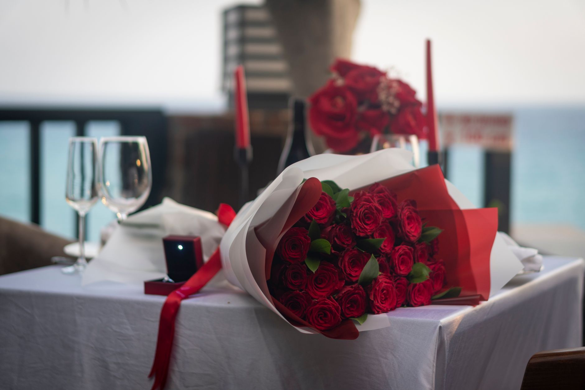 A bouquet of red roses wrapped in paper on a dinner table beside a ring box, glasses, and red candles