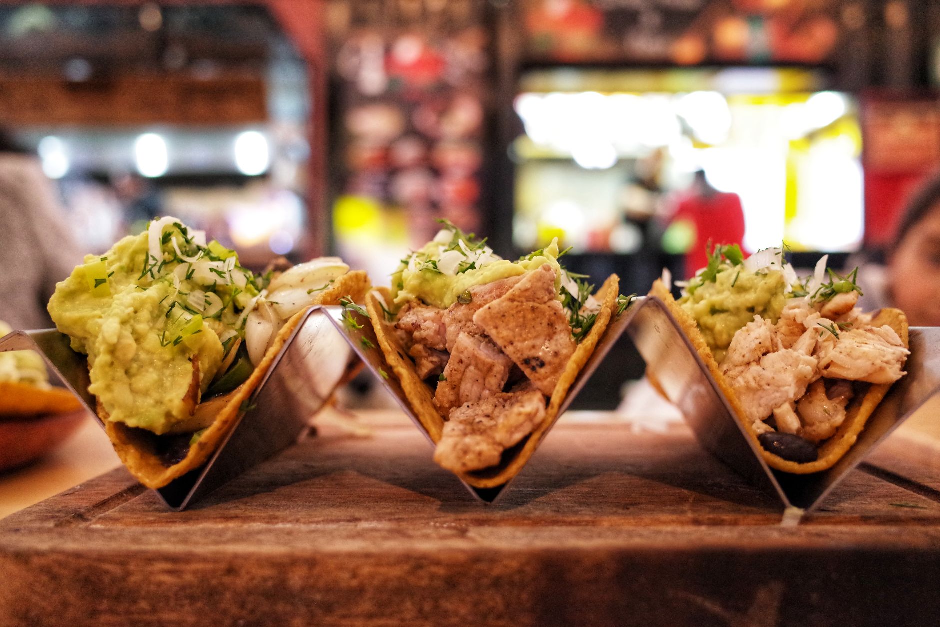 Three tacos in metal holders topped with guacamole, herbs, and onions on a wooden board