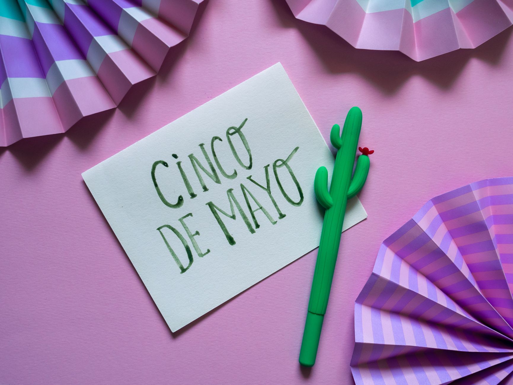 A Cinco de Mayo card, a cactus pen, and purple paper fans on a pink background