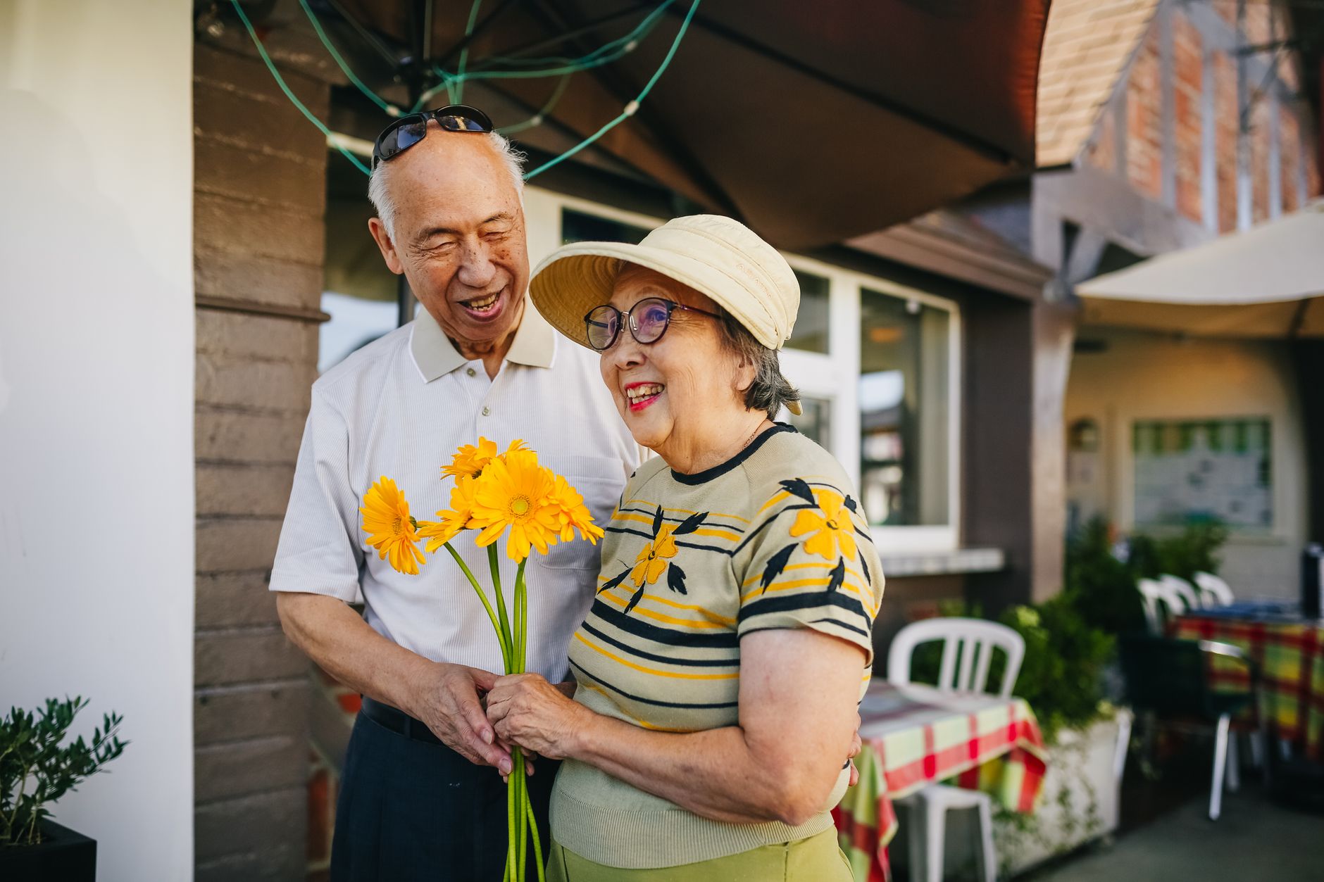 A smiling older couple holding yellow gerbera daisies outside a cafe