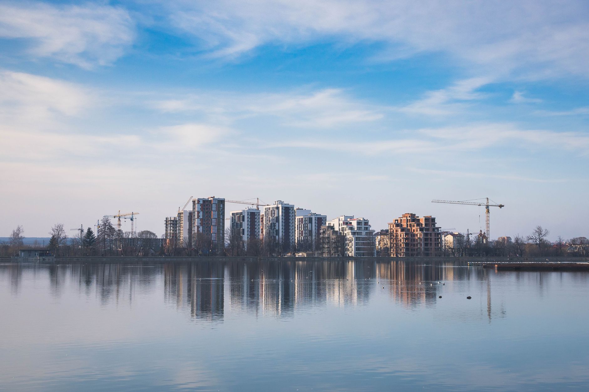 A city skyline of new apartment towers and construction cranes reflected in a calm river