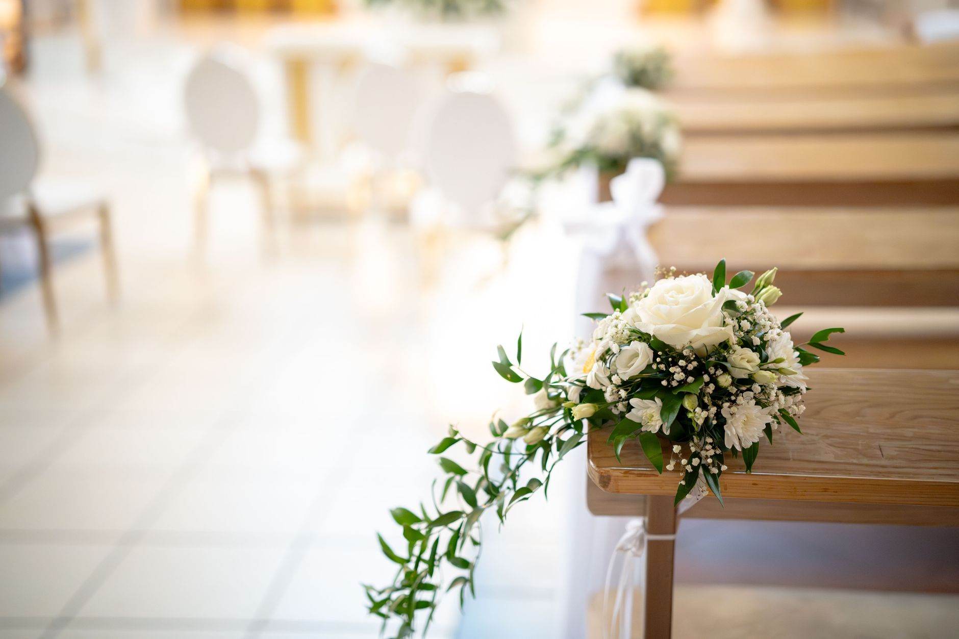A small arrangement of white roses and greenery on the end of a wooden pew at a wedding