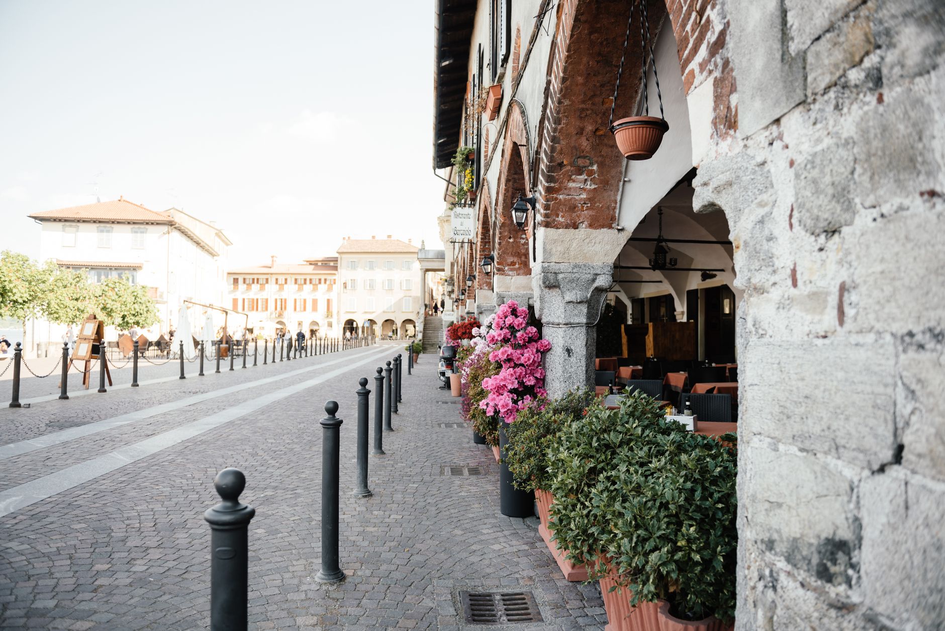 A cobblestone street with historic stone arcades, outdoor tables, and pink flowers in pots