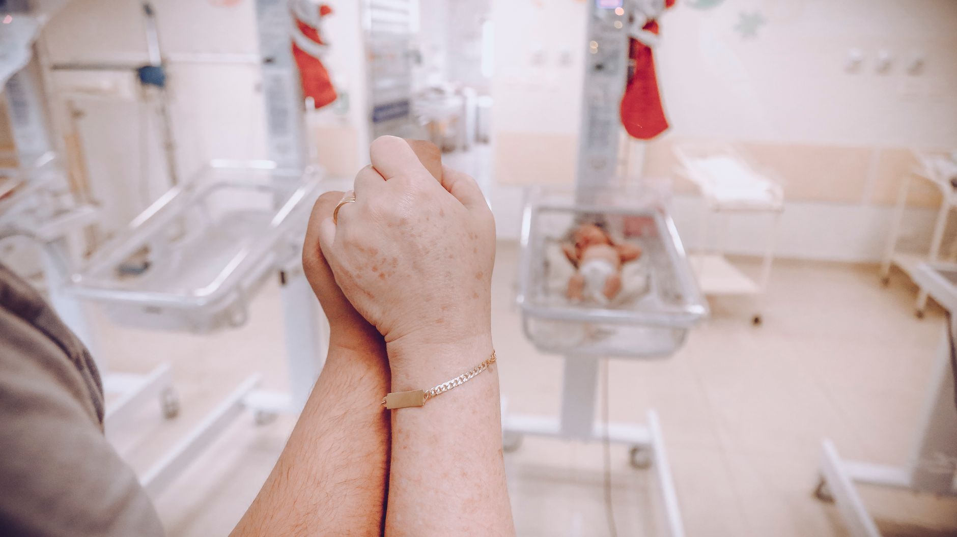 A parent&rsquo;s clasped hands in the foreground of a hospital nursery with newborns in bassinets