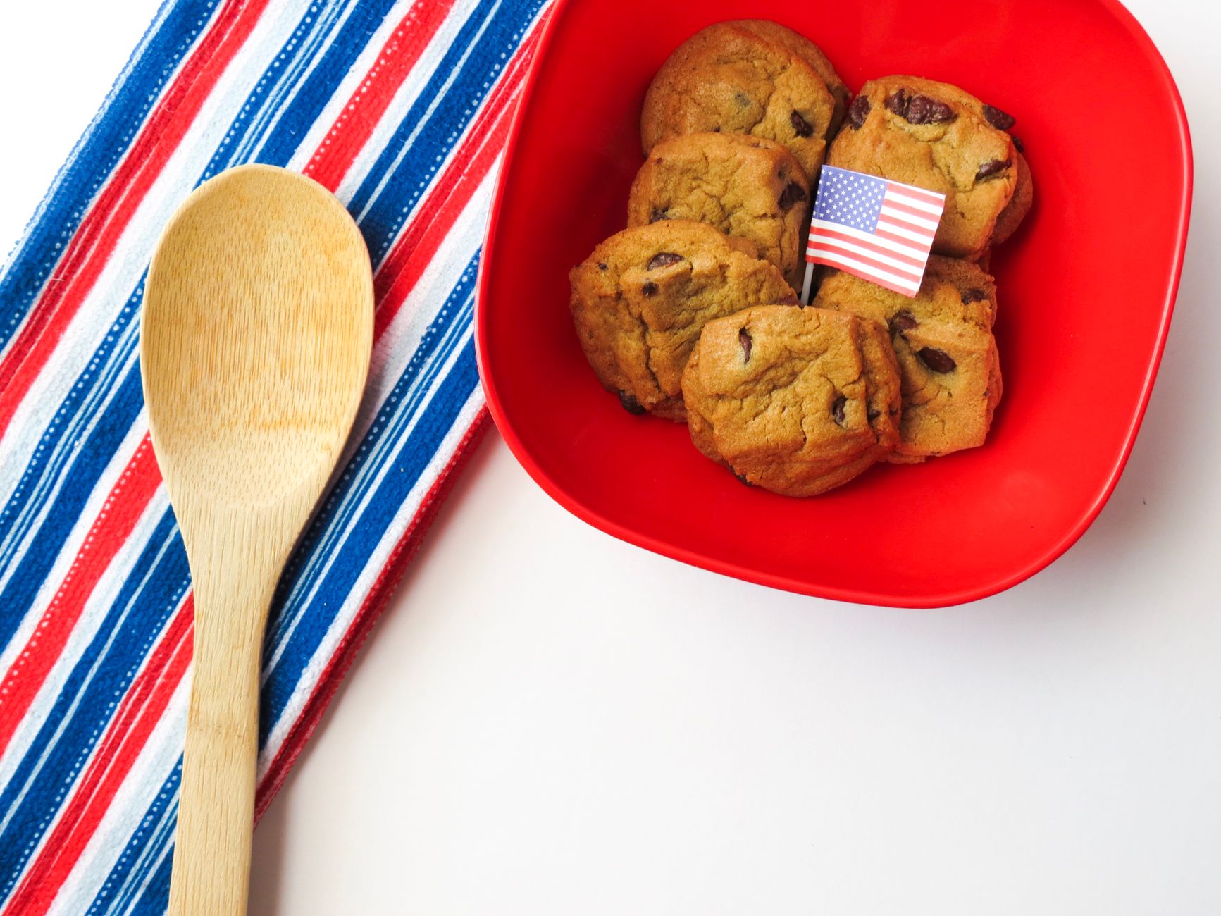 A red bowl of chocolate chip cookies with a small American flag next to a striped red, white, and blue towel