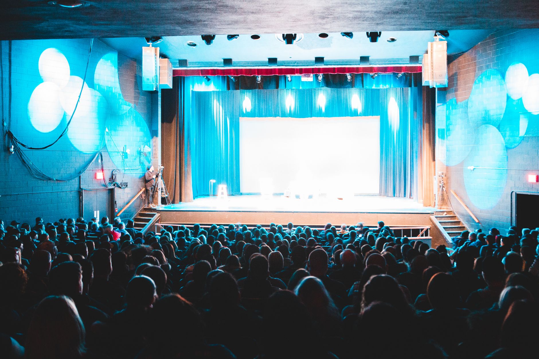 An audience watching a lit stage in a theater with a blue curtain