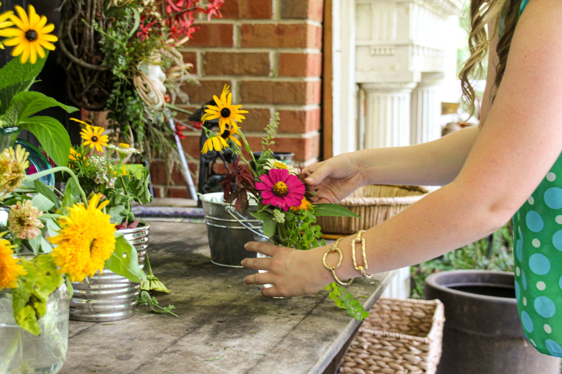 A woman arranging a pink zinnia and yellow flowers in a metal bucket on a garden table