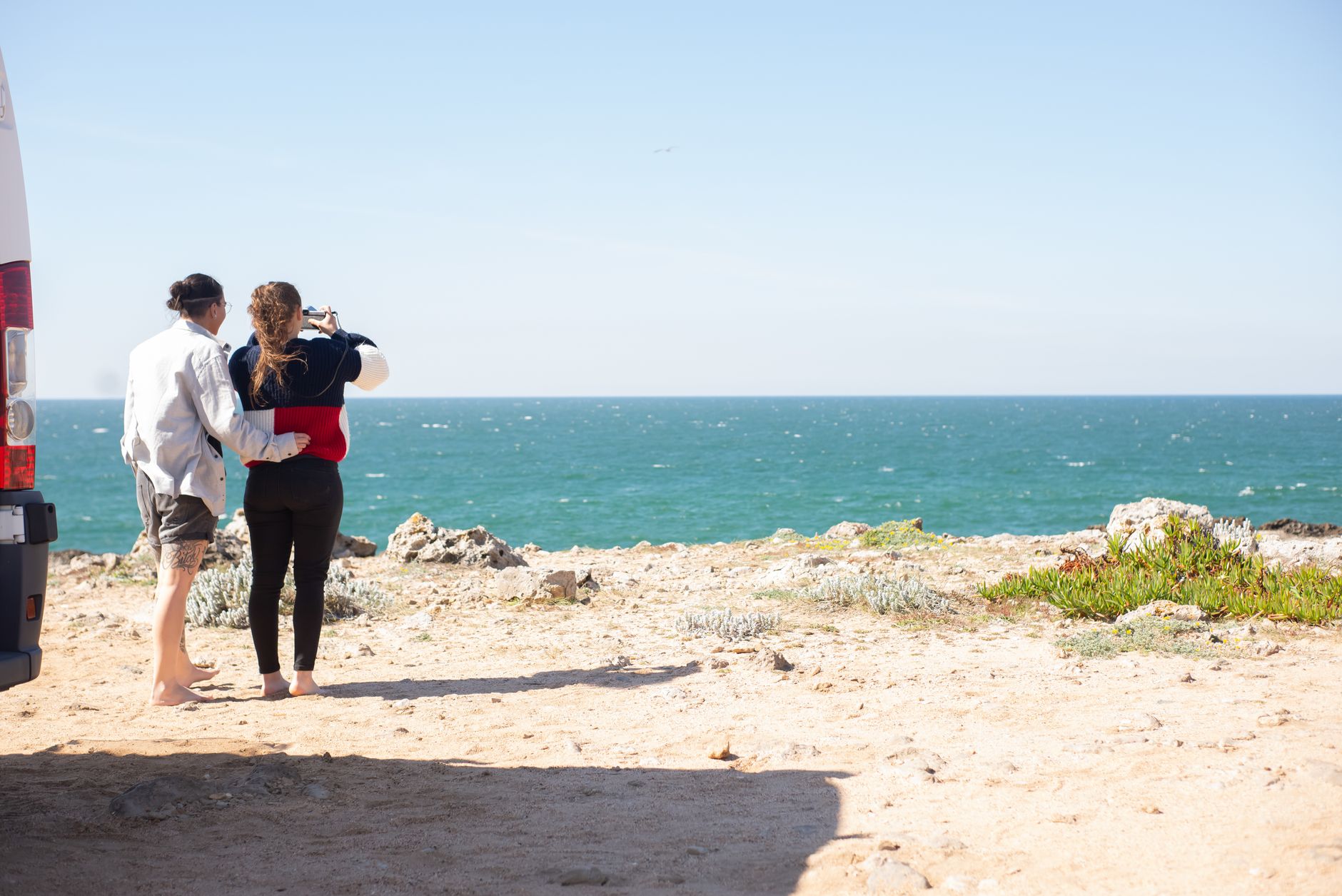 Two people standing beside a camper van looking at the ocean from a sandy bluff