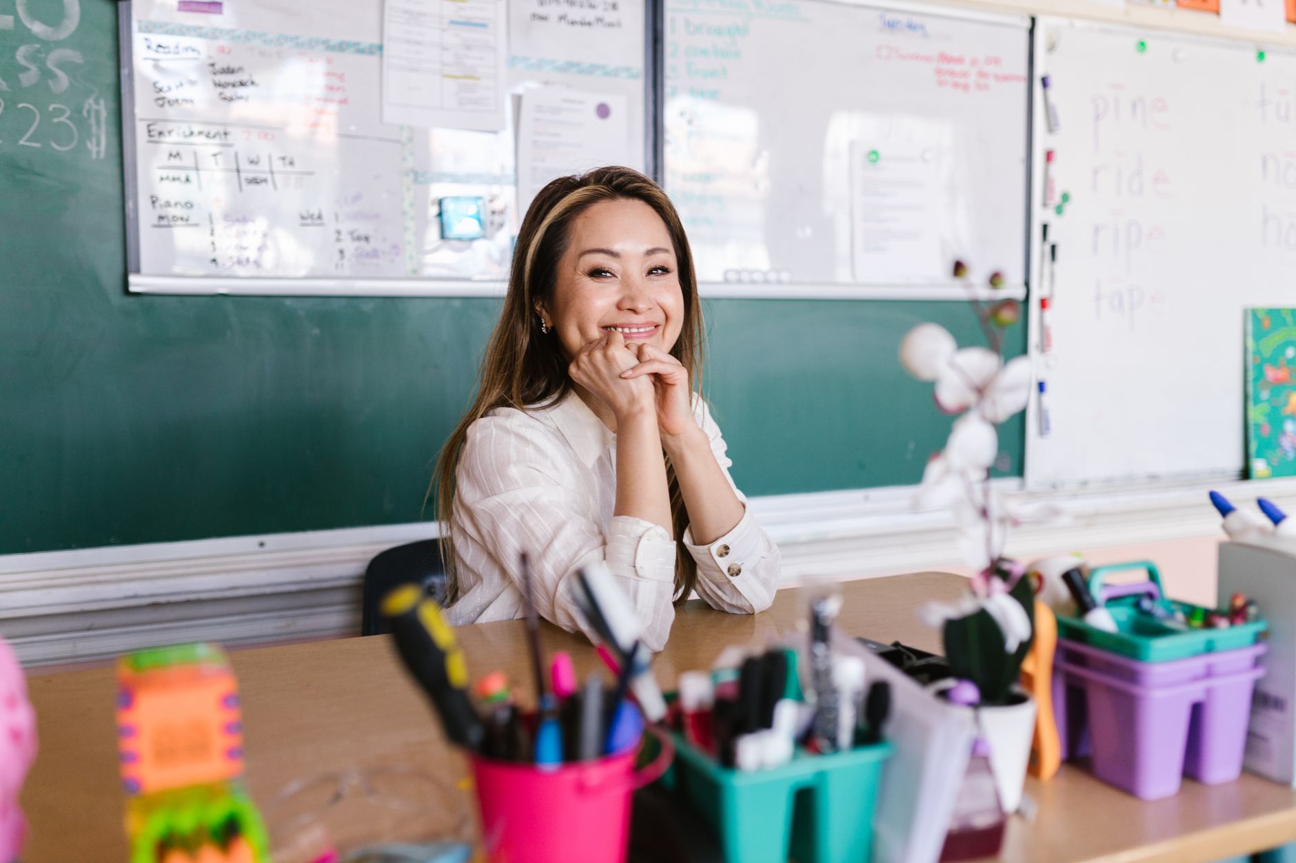 A smiling teacher seated at a desk with pencil cups in front of a classroom whiteboard