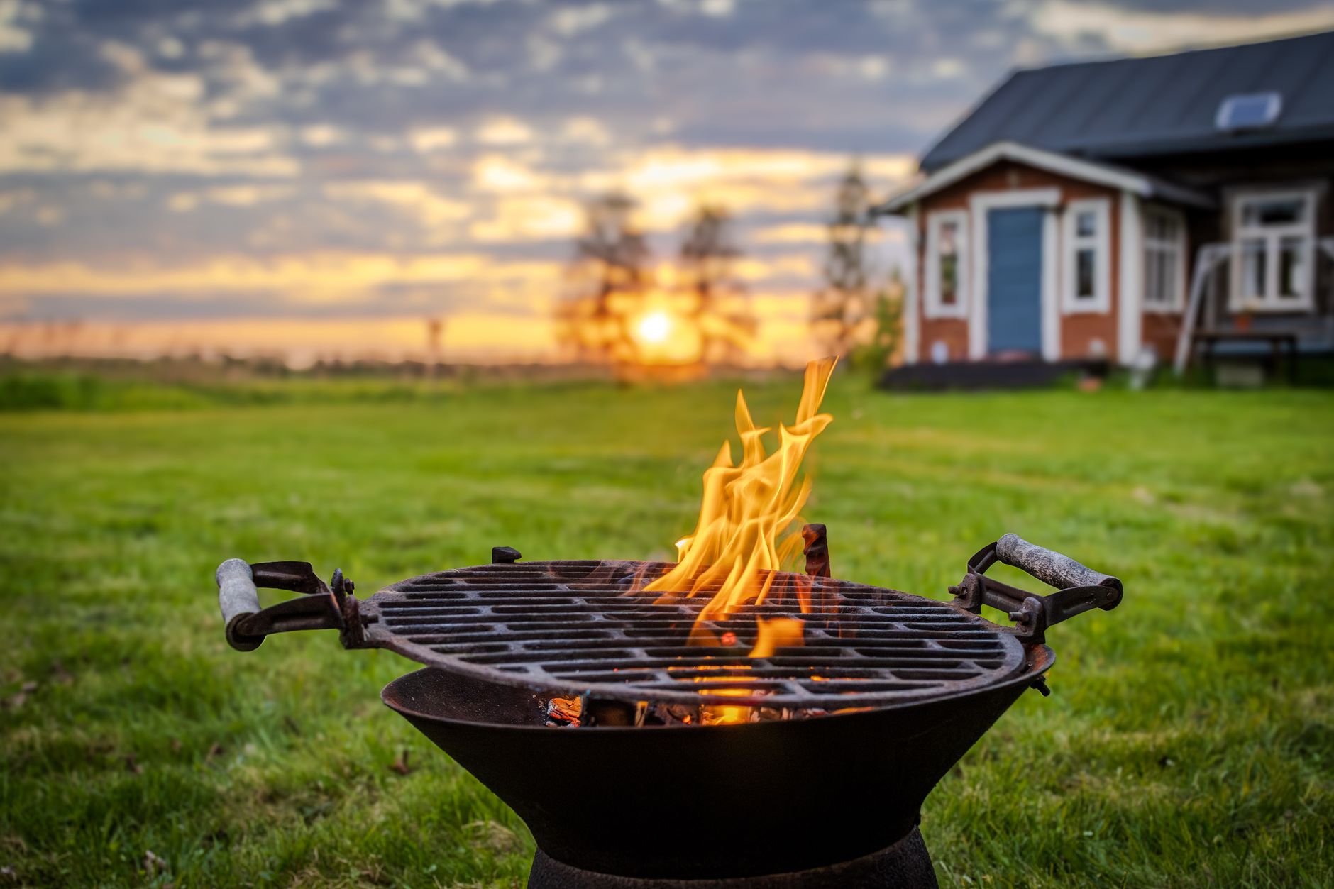 A flaming charcoal grill on a lawn in front of a red cabin at sunset