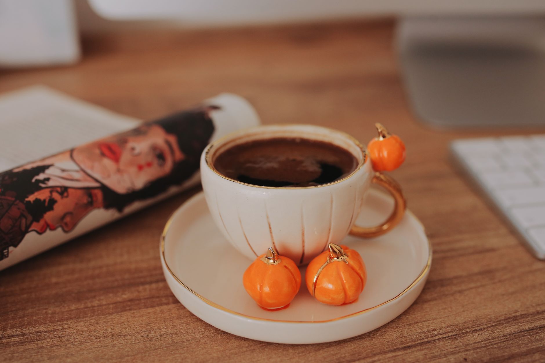 A cup of black coffee with small orange pumpkin charms on a saucer beside a keyboard and a magazine