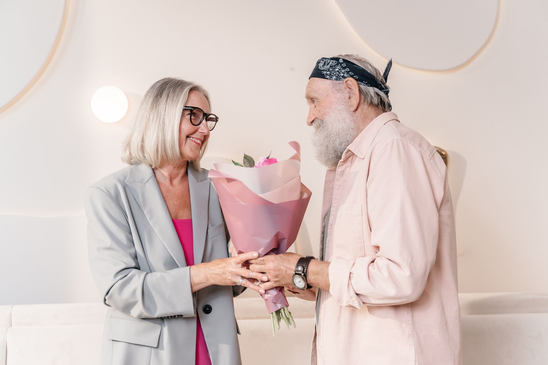 An older man handing a wrapped bouquet of pink flowers to a smiling older woman