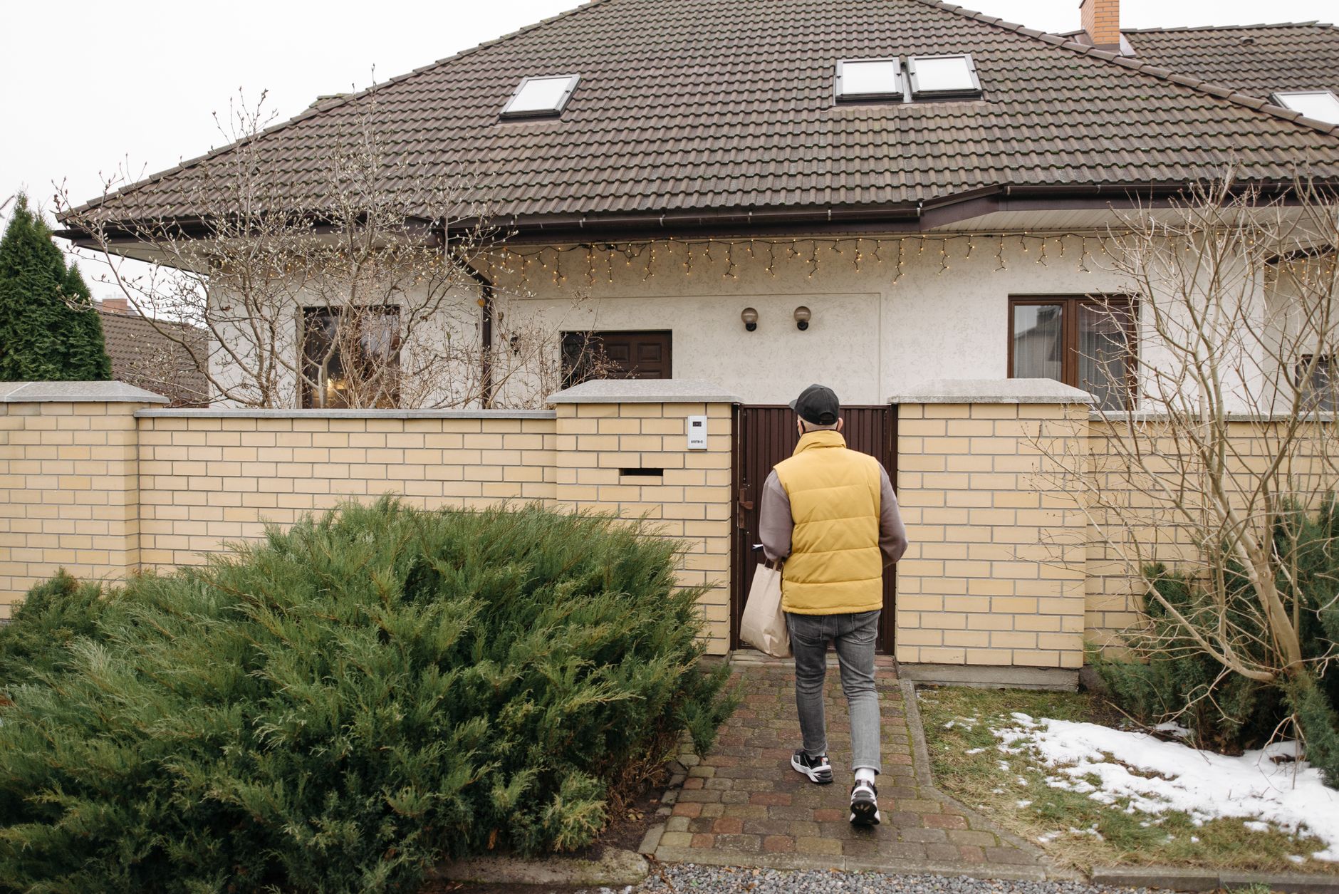 A delivery person in a yellow vest carrying a bag toward the gate of a house