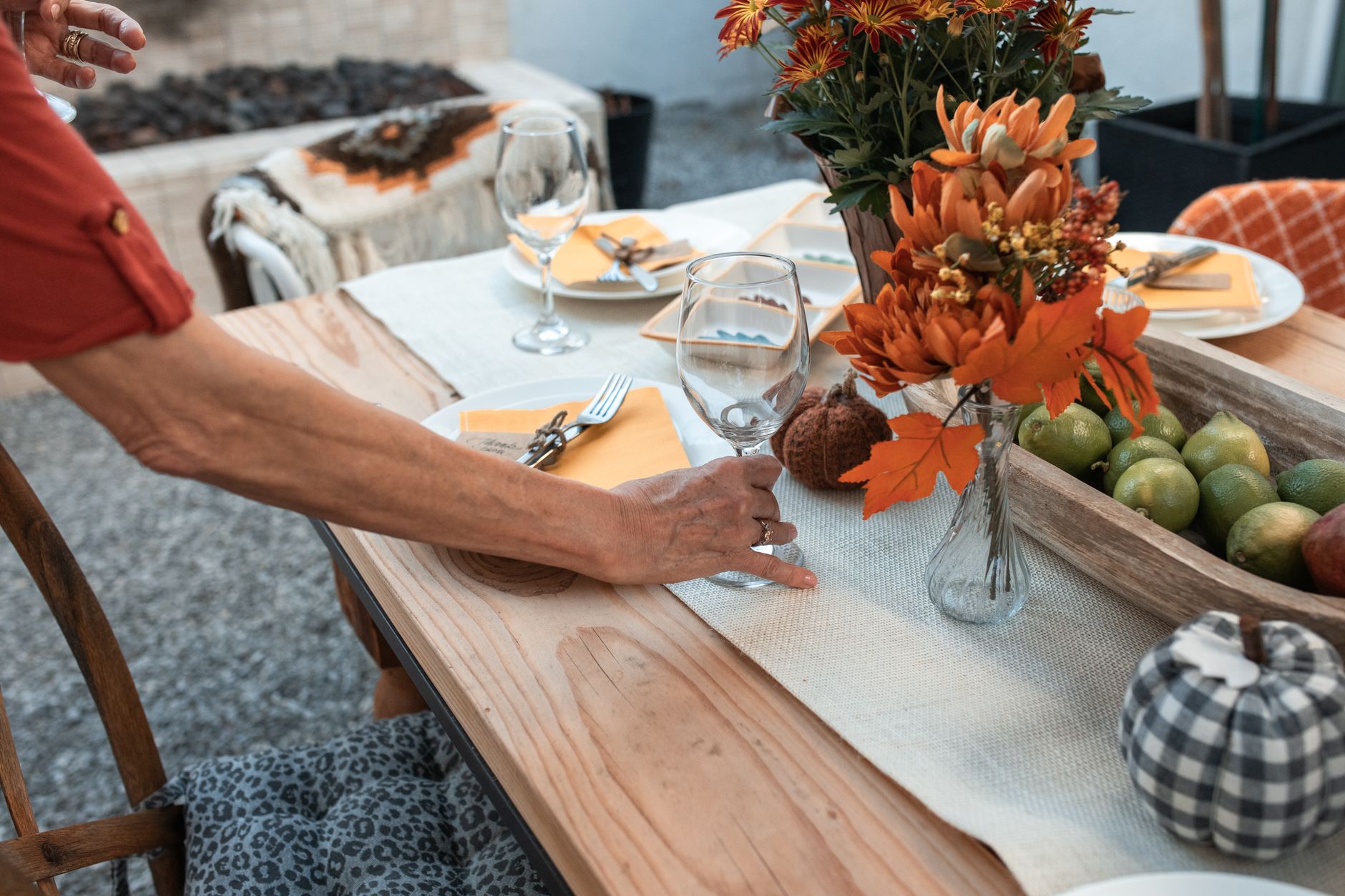 A person setting a fall dinner table with orange flowers in a small vase, pears, and a checked pumpkin