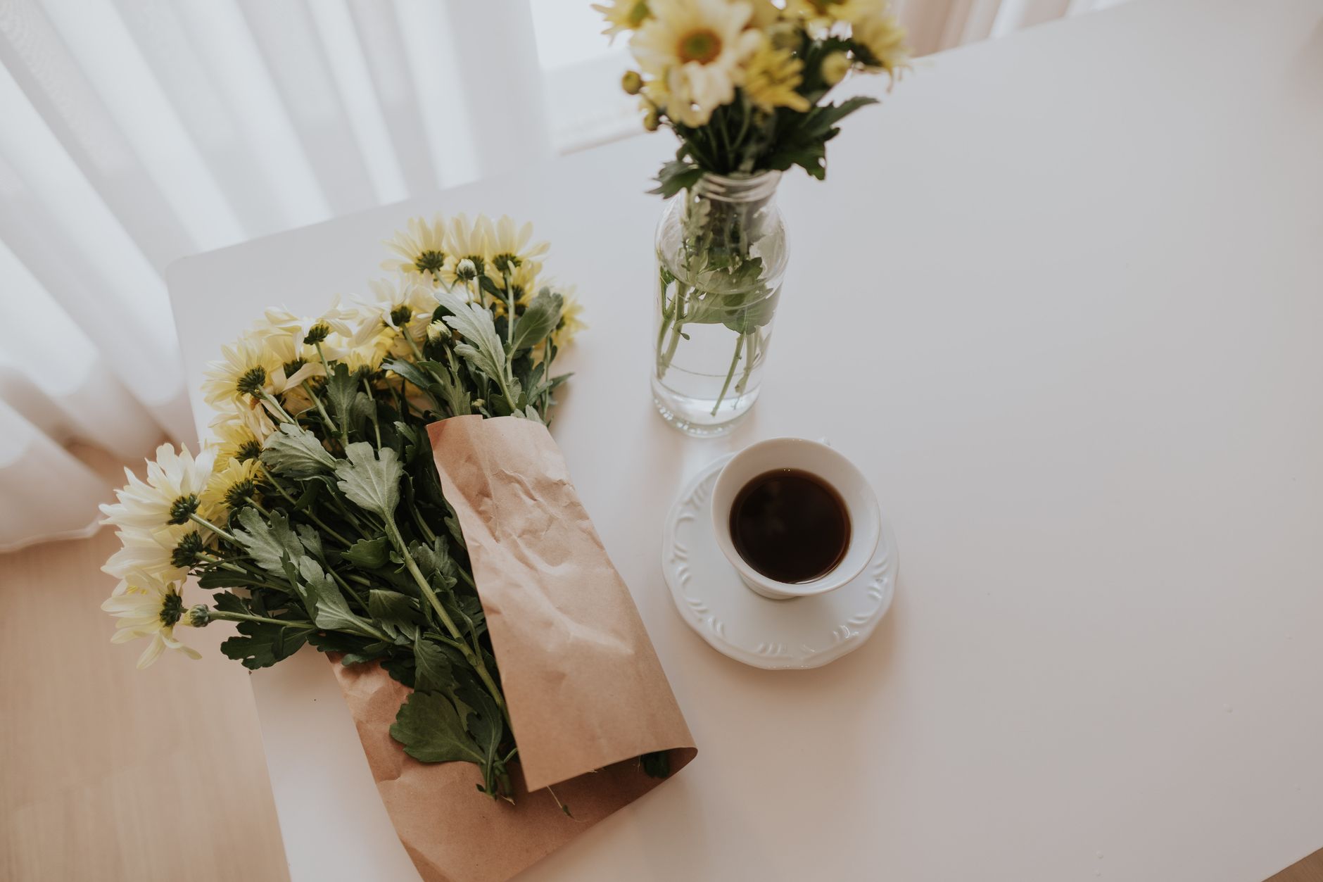A bunch of yellow and white chrysanthemums in brown paper on a white table beside a glass jar of the same flowers and a cup of coffee