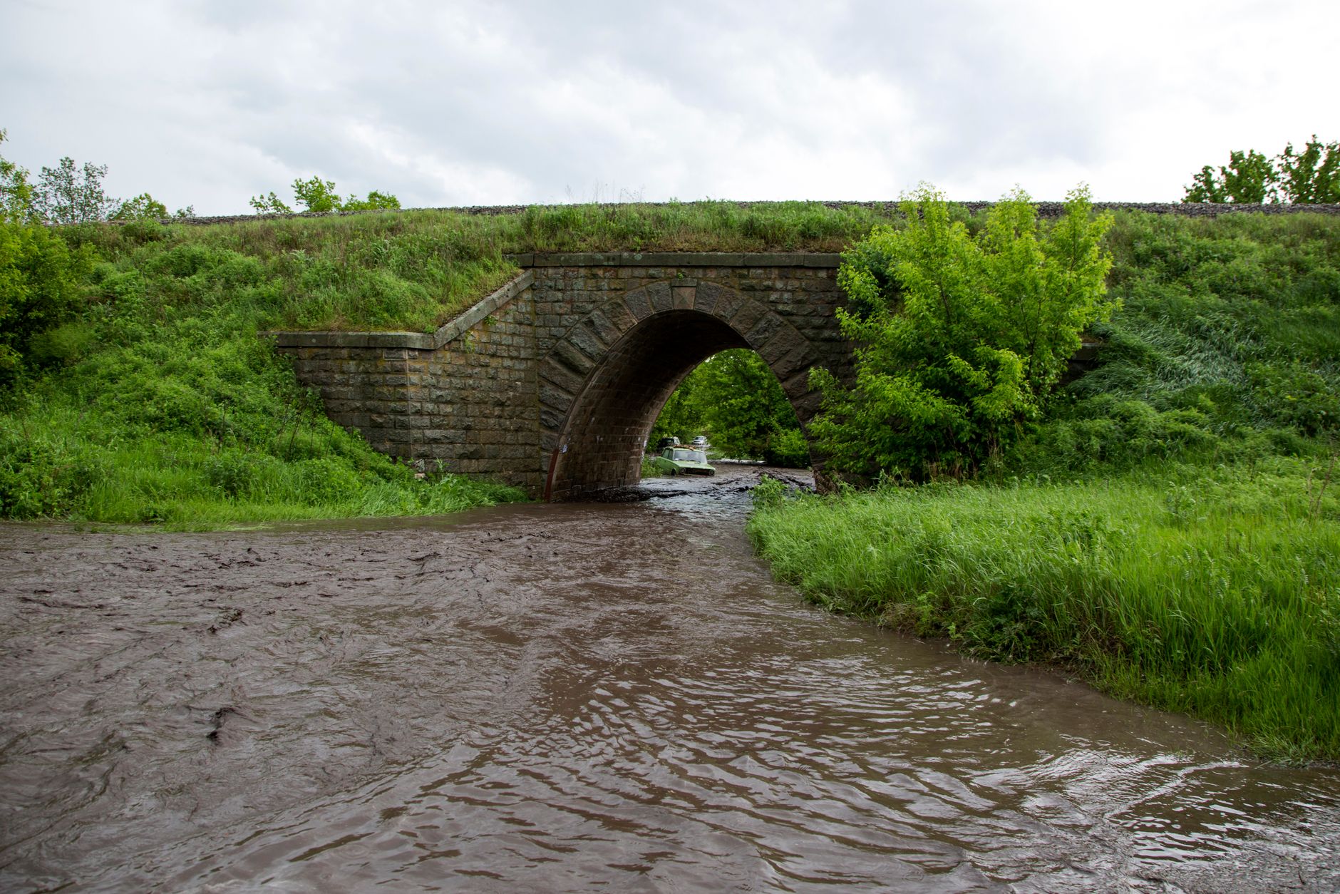 An old stone arch bridge over a muddy creek running through green fields