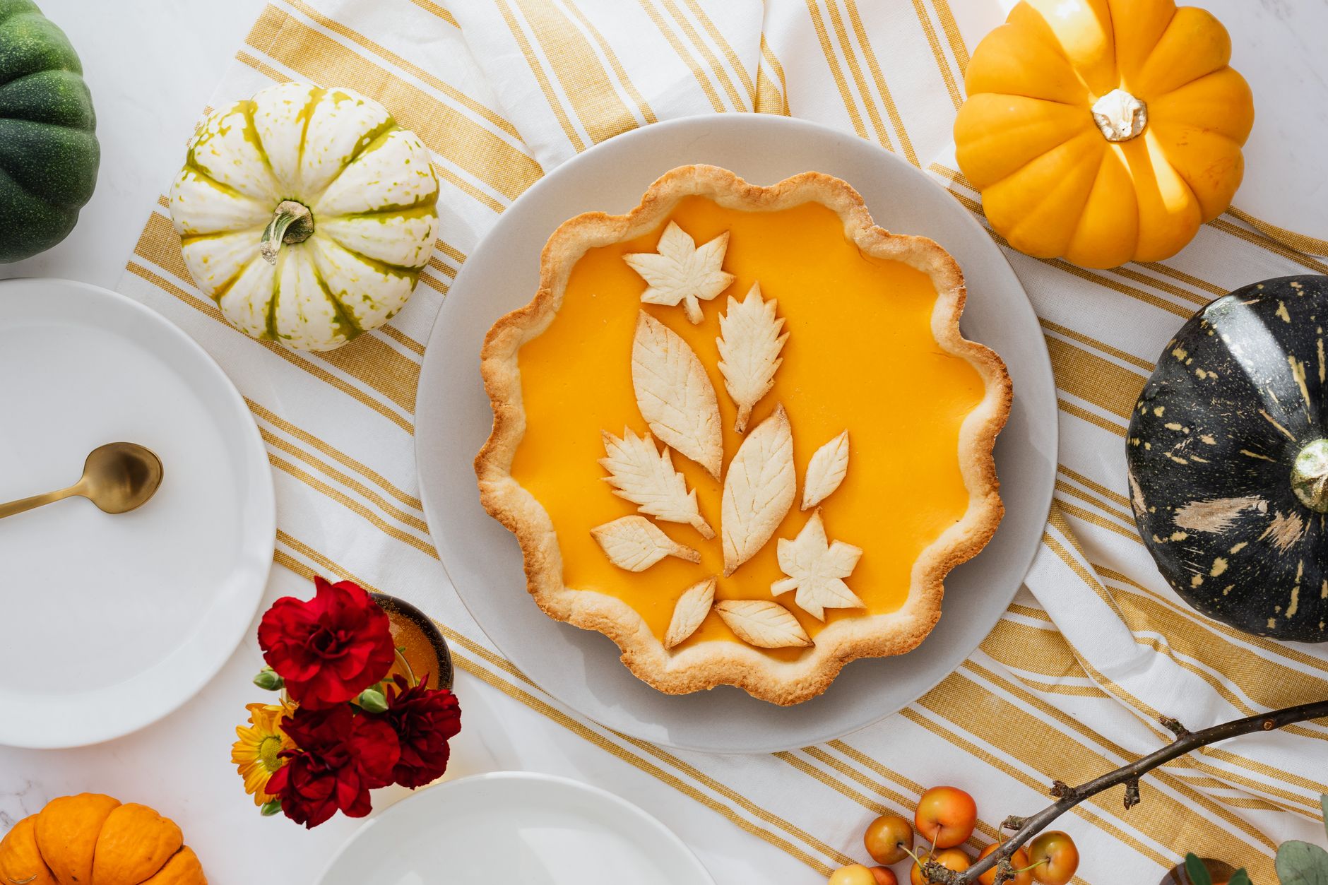 A pumpkin pie with leaf-shaped pastry cutouts on a striped table, surrounded by small pumpkins and a small vase of red flowers